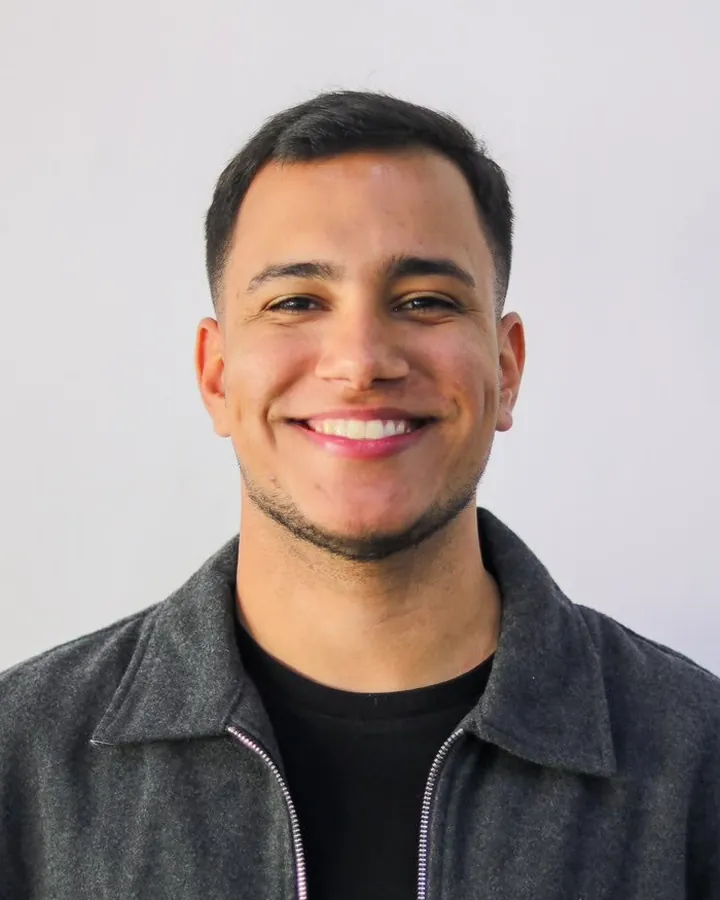 Foto de Lucas Ricardo sorrindo, de jaqueta cinza, num fundo claro